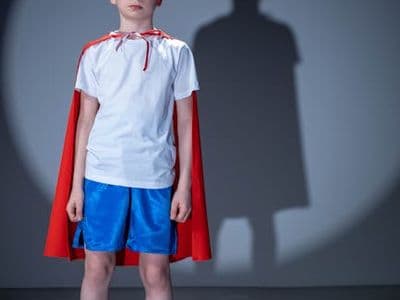 A child dressed as a superhero, wearing a red cape and mask, standing in spotlight shadow.
